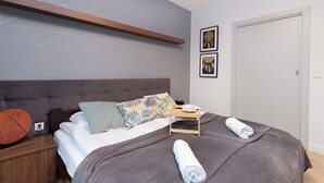 A modern bedroom with a large bed, a bedside table, a nightstand, a wooden shelf, and two framed pictures on the wall.