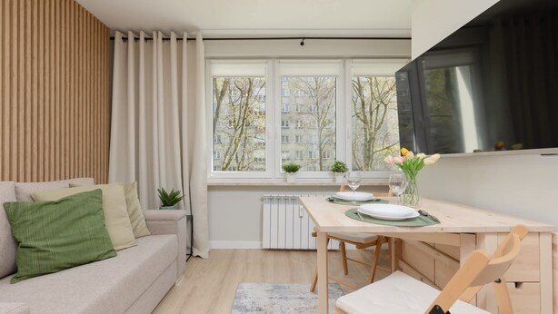 Una sala de estar moderna con sofá, una mesa de comedor con sillas, televisor montado en la pared y ventana con vista a edificios y árboles.