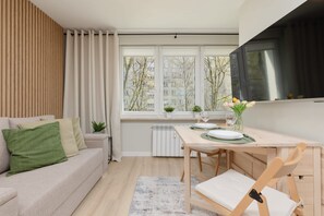 A modern living room with a sofa, a dining table with chairs, a TV mounted on the wall, and a window with a view of buildings and trees. - Cozy Studio in Downtown | POLIN Museum (Warszawa)
