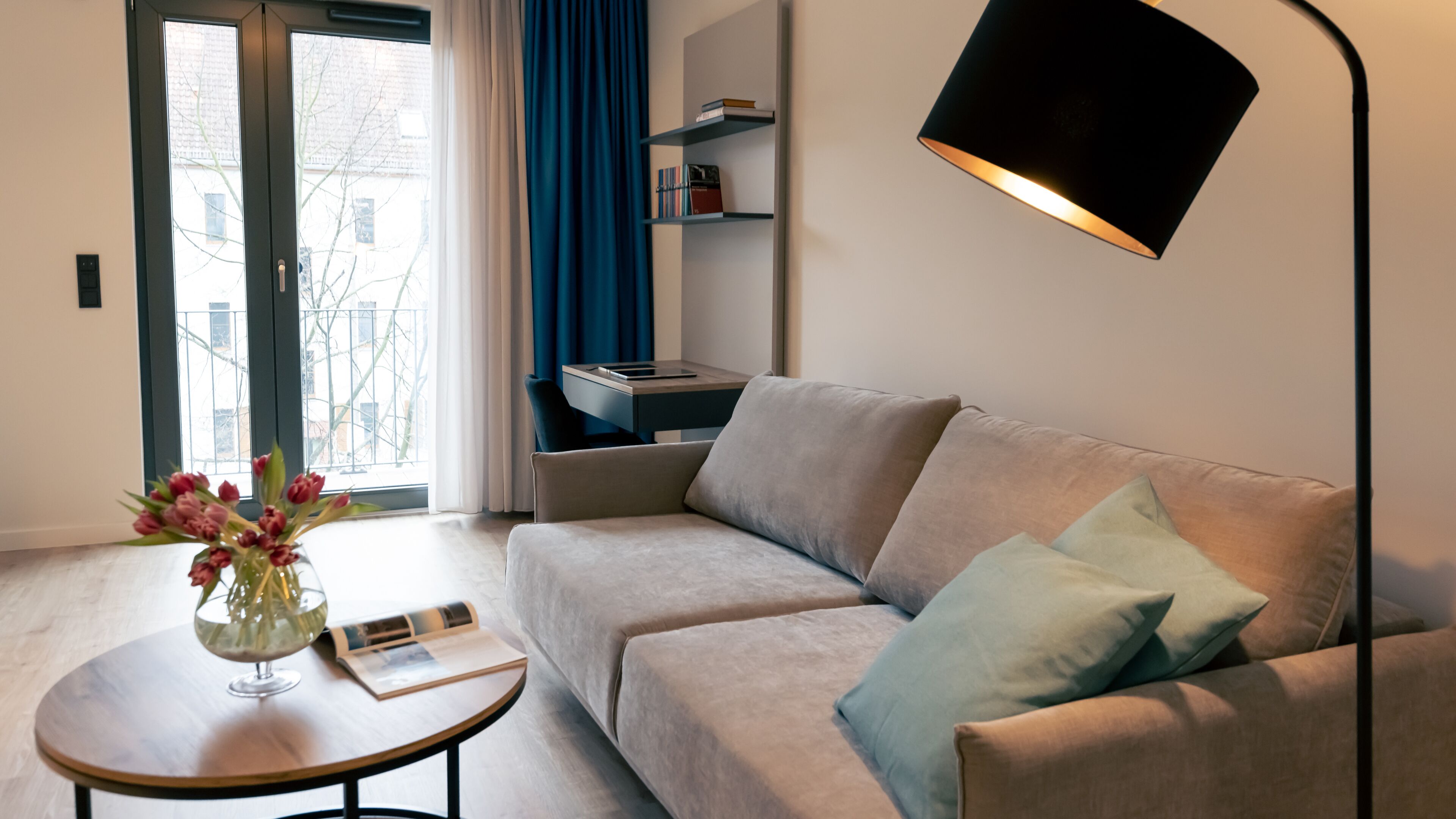 A modern living room with a sofa, a coffee table with a vase of flowers, a bookshelf, and a floor lamp.