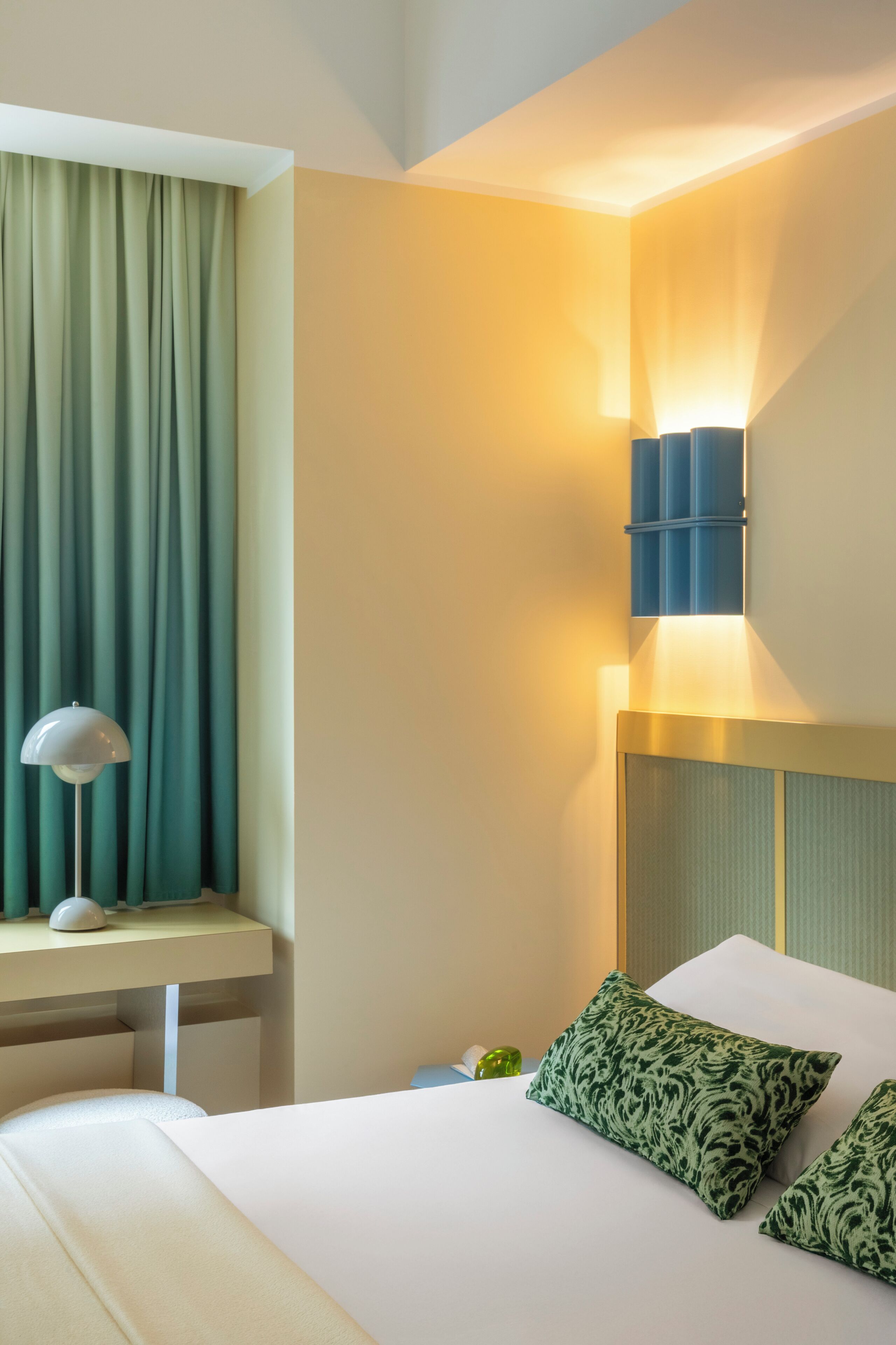 Une chambre d’hôtel avec un lit, un bureau, une lampe, un luminaire fixé au mur et un rideau.