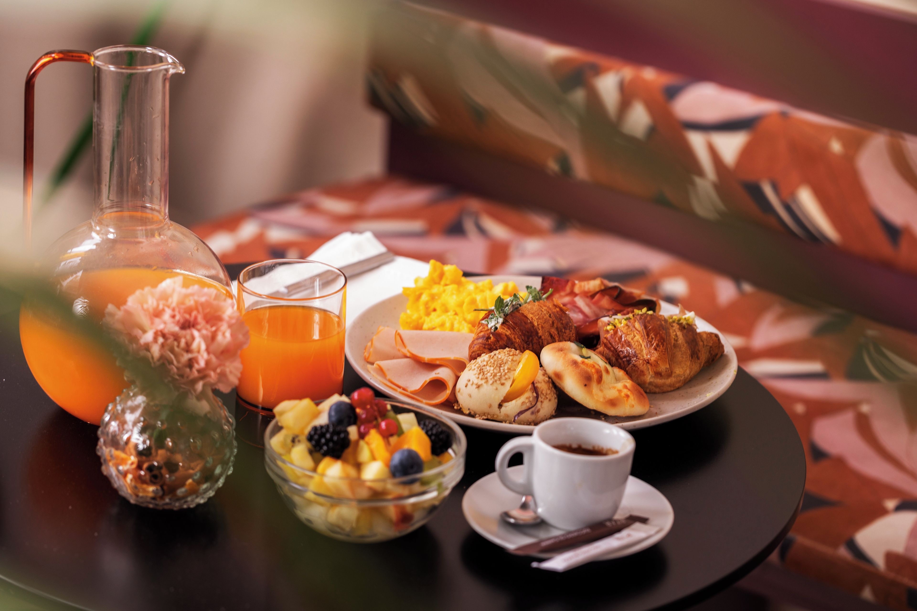 A breakfast table with a pitcher, glasses, a bowl of fruit, a plate with eggs, ham, and pastries, and a cup of coffee.
