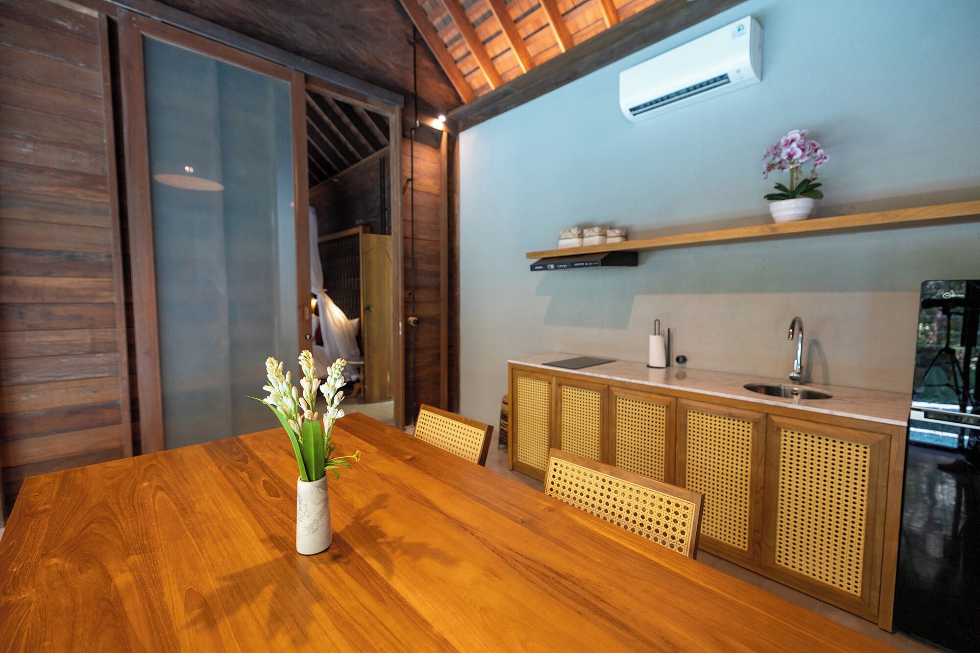 A wooden dining table with a vase of flowers, a kitchen with a sink and cabinets, and an air conditioning unit on the wall.