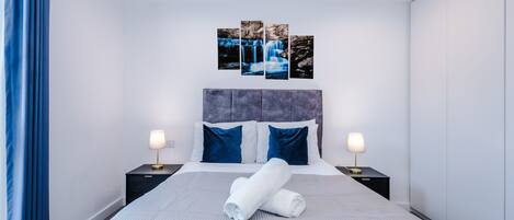 A modern hotel room with a large bed, two bedside tables, a headboard, and a wall-mounted artwork.