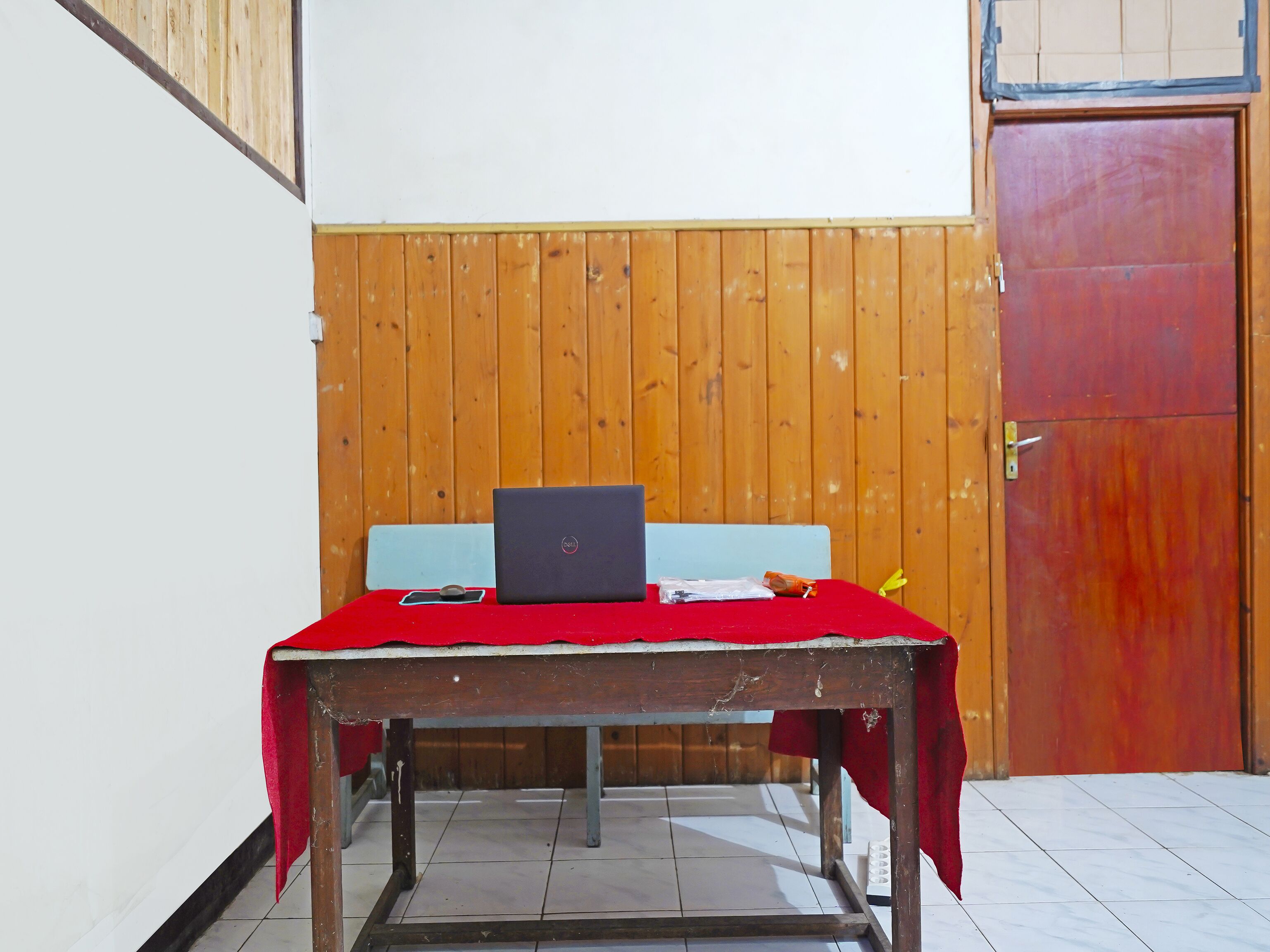 Una habitación pequeña con una puerta de madera, una mesa roja con un ordenador portátil y una silla azul.
