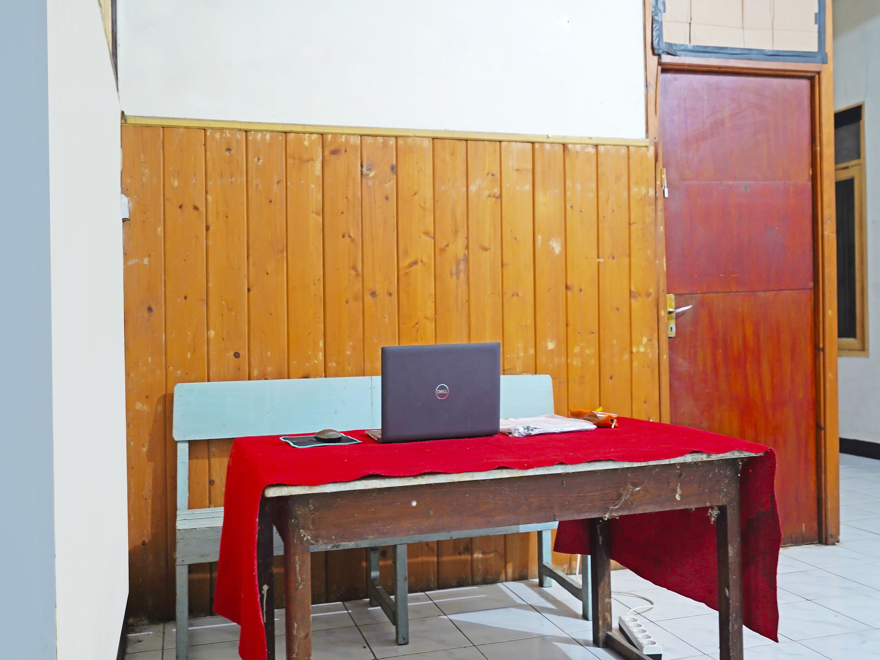 Una habitación pequeña con un banco de madera, una mesa con una computadora portátil y un paño rojo.