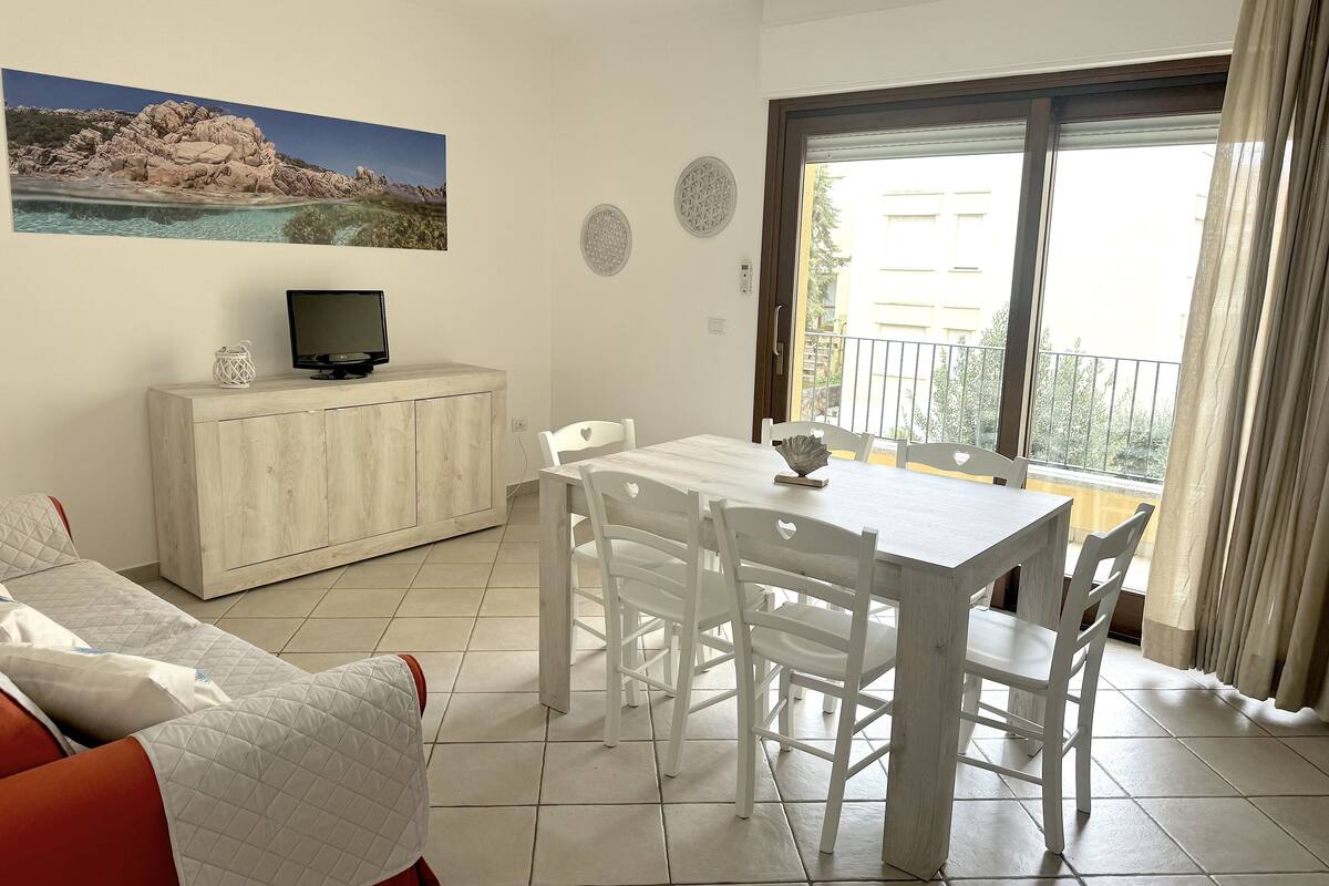 A dining area with a table and chairs, a TV on a cabinet, and a balcony with a view.