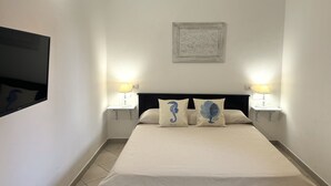 En pænt redt seng med et beige sengetæppe og to dekorative puder, med to sengeborde og lamper på hver side.