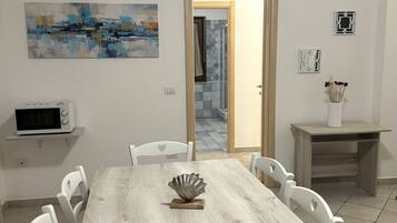 Un comedor con una mesa de madera, sillas blancas, un microondas en un estante, un centro de mesa decorativo con conchas marinas y un cuadro en la pared.