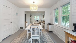 A dining area with a white table, chairs, and a chandelier. There is a window with a view of trees.