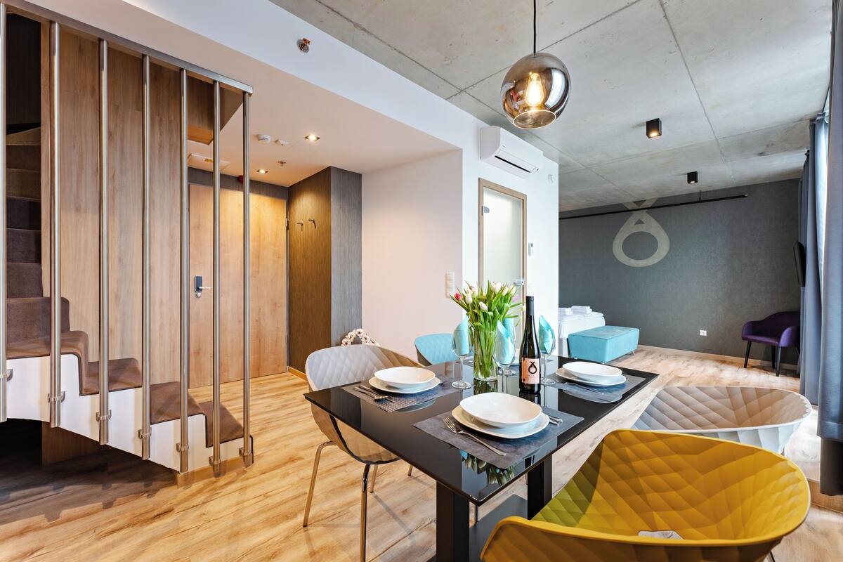 A modern dining area with a black table, white plates, and a bottle of wine. There are chairs and a yellow ottoman.