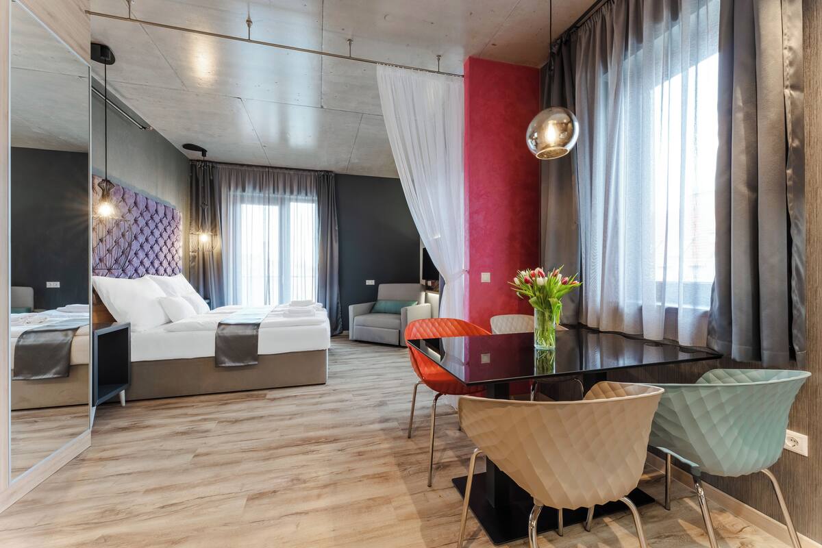 A modern hotel room with a bed, a dining table with chairs, a mirror, and a large window with curtains.
