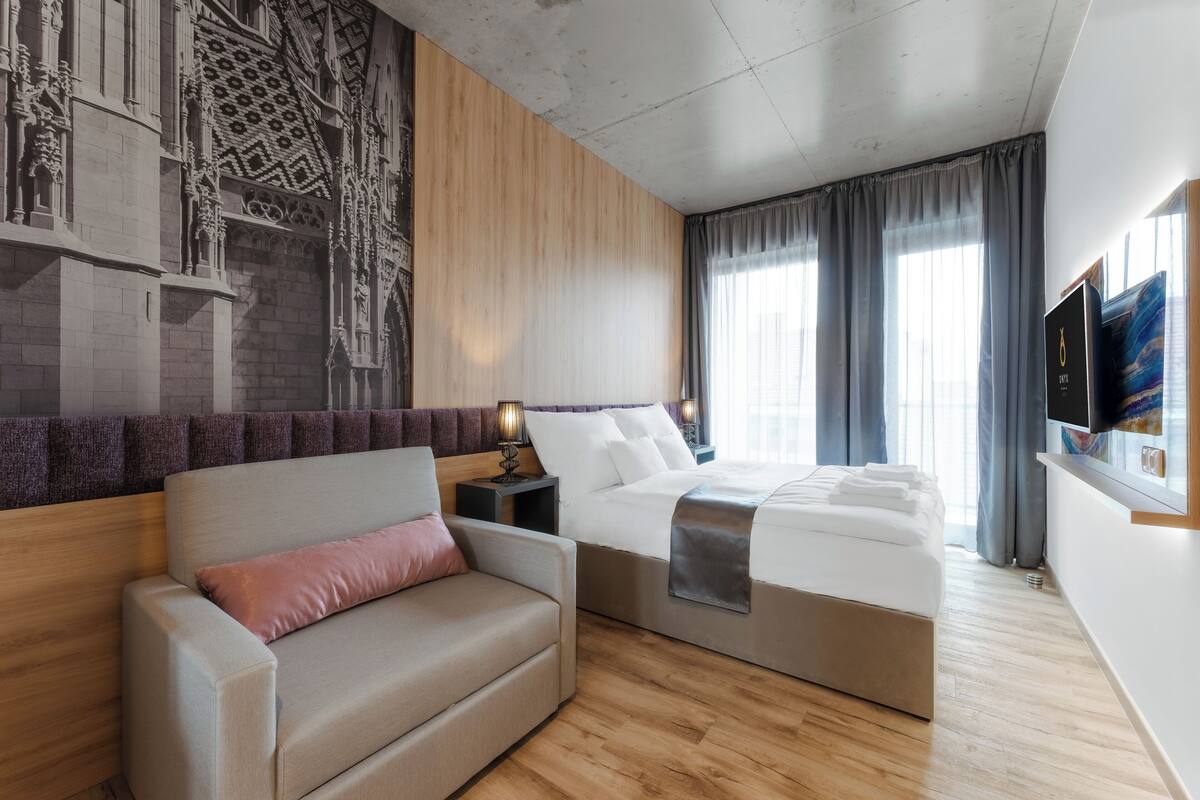 A modern hotel room with a wooden accent wall, a large bed, a sofa, and a flat-screen TV.