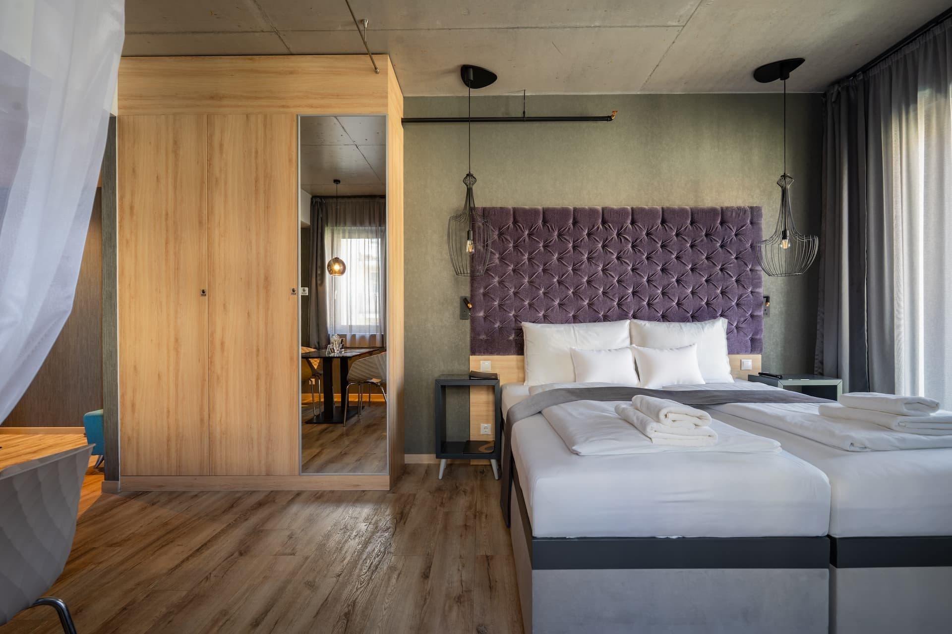 Une chambre d’hôtel moderne dotée d’un grand lit, d’une armoire en bois, d’un bureau avec une chaise et offrant une vue sur la salle à manger.