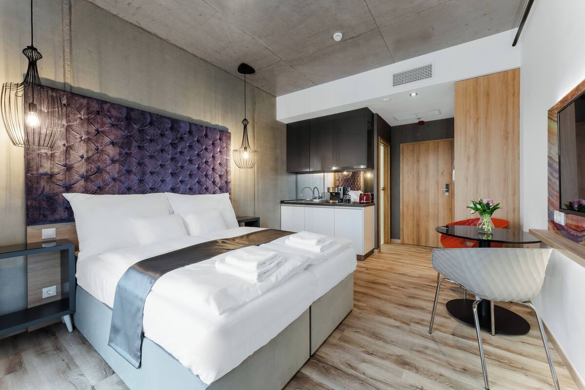 A modern hotel room with a large bed, a built-in desk, a kitchenette, and a dining area.