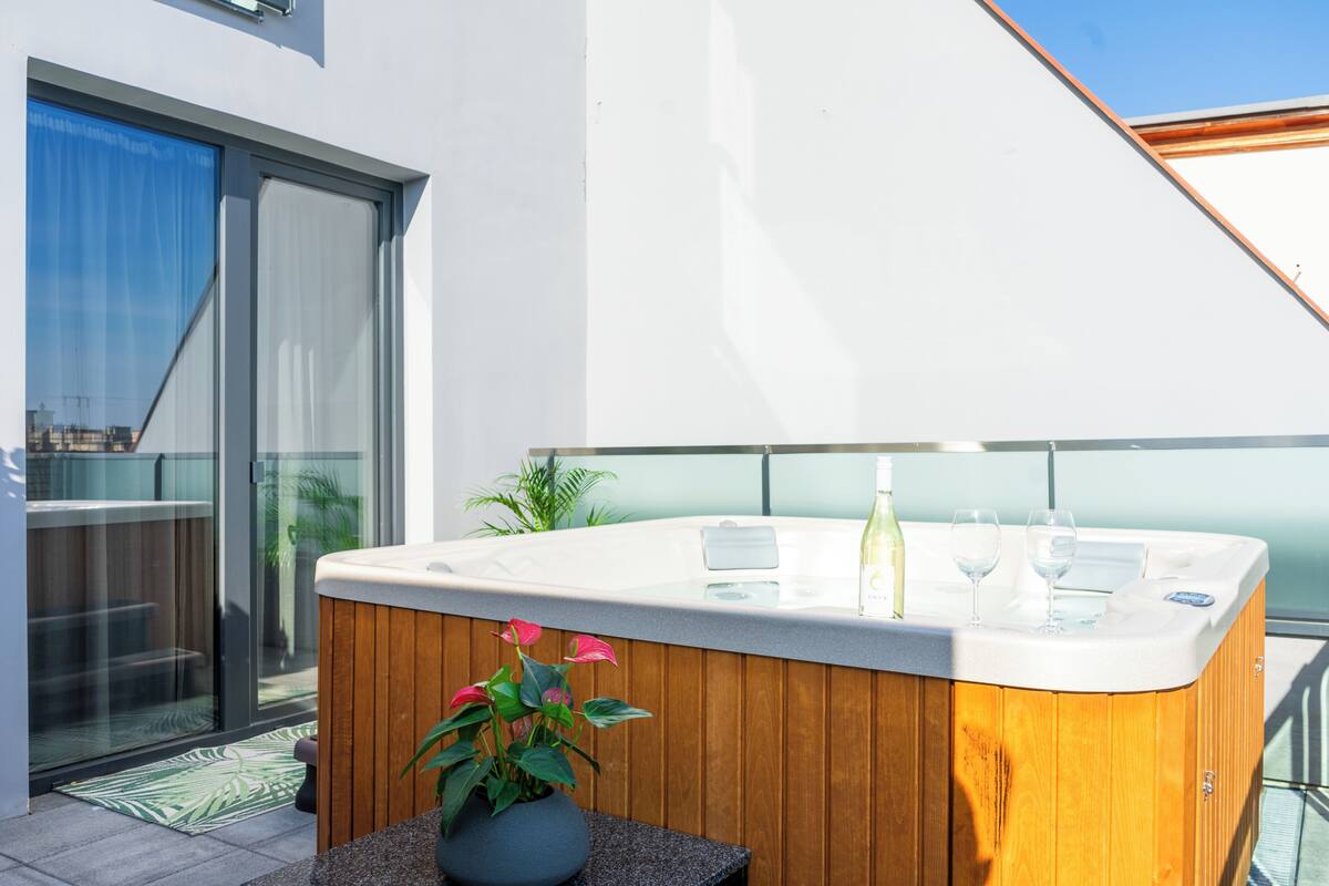 A modern outdoor hot tub with a wooden exterior, a glass door leading to a balcony, and a potted plant nearby.