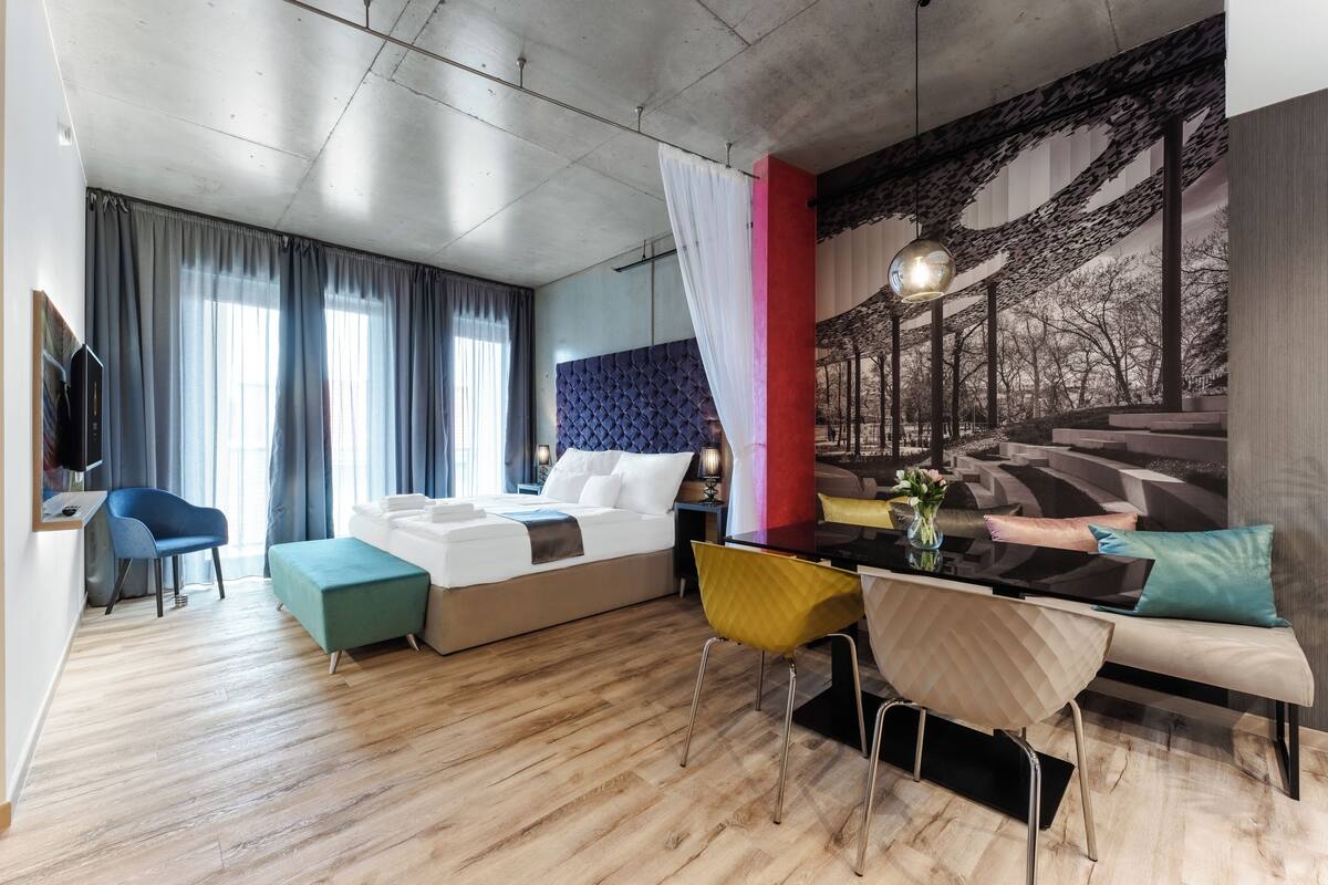A modern hotel room with a bed, a dining table, and a large mural on the wall.