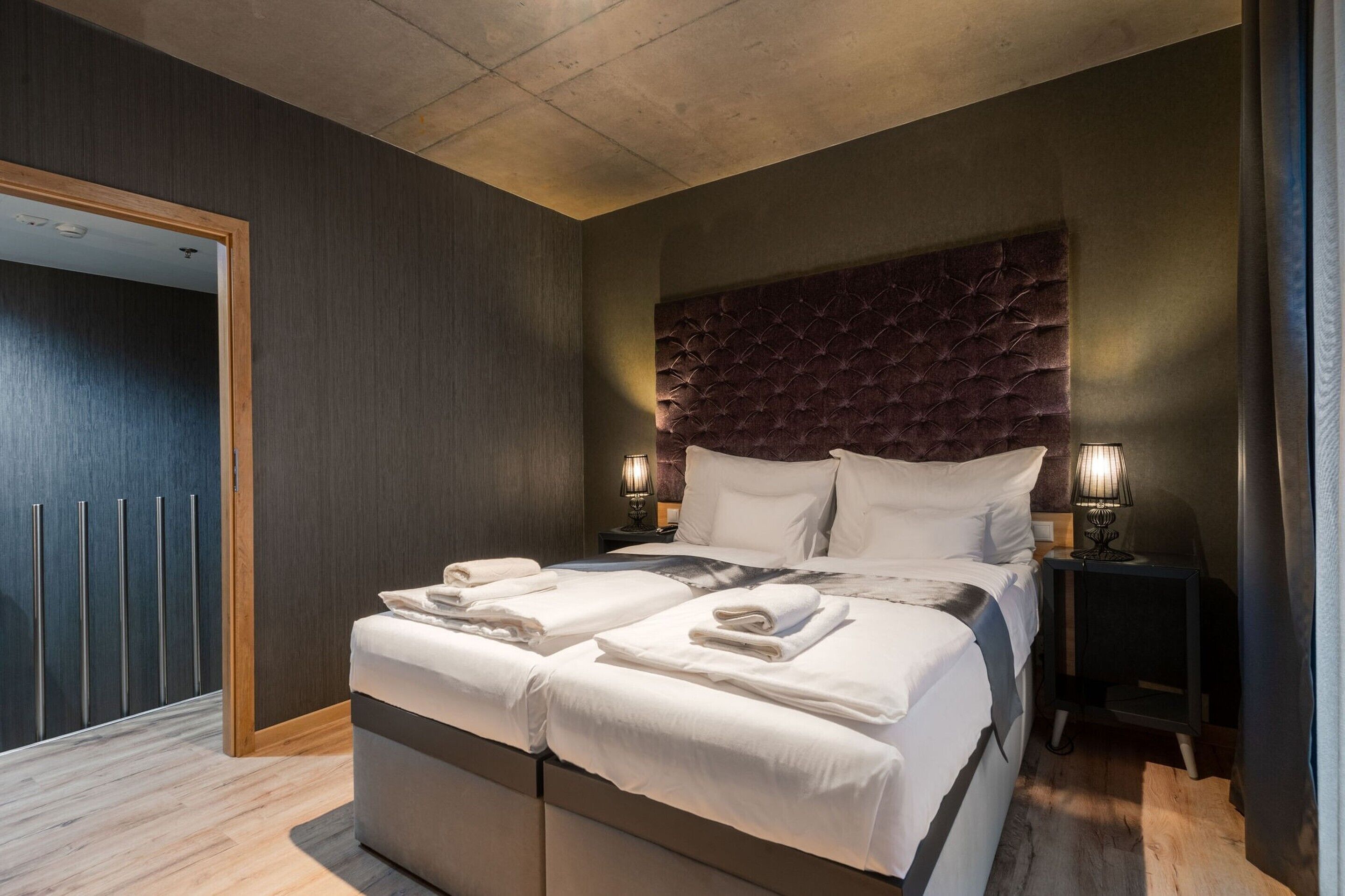 Een moderne hotelkamer met een groot bed, twee nachtkastjes met lampen, een donker hoofdeinde en een houten garderobe.