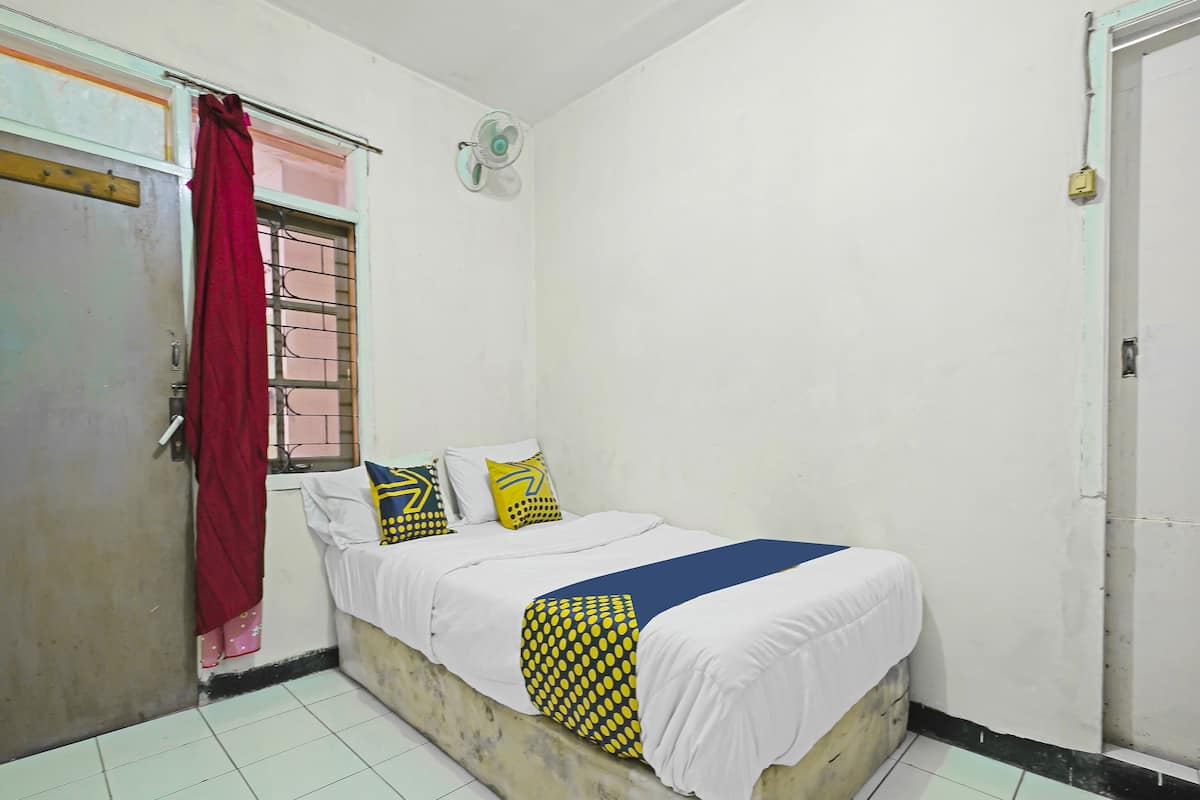 A small, single-bed room with a white bedspread, two pillows, a red curtain, a window with bars, a wall-mounted fan, and a door.