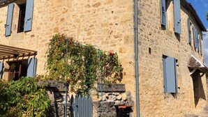A stone building with a tiled roof, a chimney, and a small garden area with a tree and a bush.