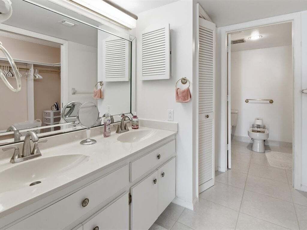 A bathroom with a double sink, a mirror, a towel rack, and a toilet.