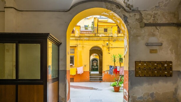 Un portail en arc donne accès à une cour intérieure où se trouvent un bâtiment jaune, un pot de fleurs et une boîte aux lettres.