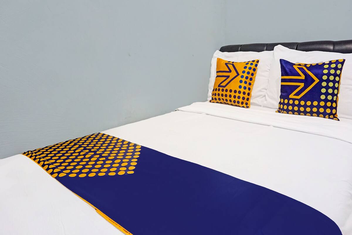 Una cama con colcha blanca y azul, dos almohadas con diseños amarillo y azul, y un cabecero oscuro.