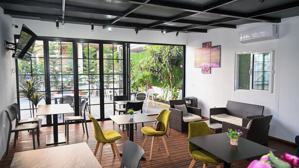 An indoor seating area with tables and chairs, large windows, and a wall-mounted TV. - VILA AARON (Durrës)