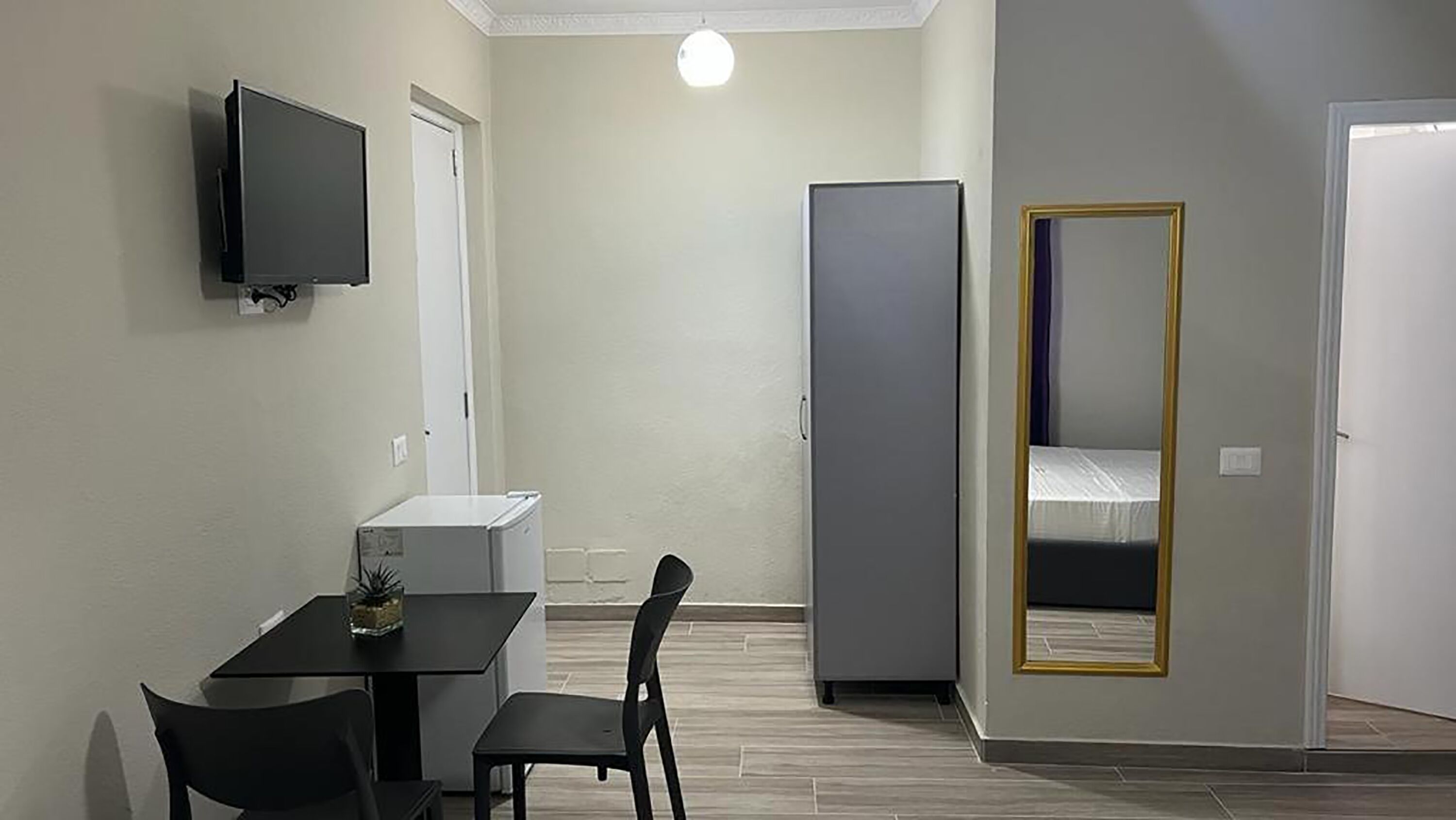 A compact room with a desk, chairs, a television, a wardrobe, and a mirror.
