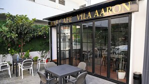 An outdoor seating area with tables and chairs, a potted plant, and a building entrance with a glass door and signage. - VILA AARON (Durrës)
