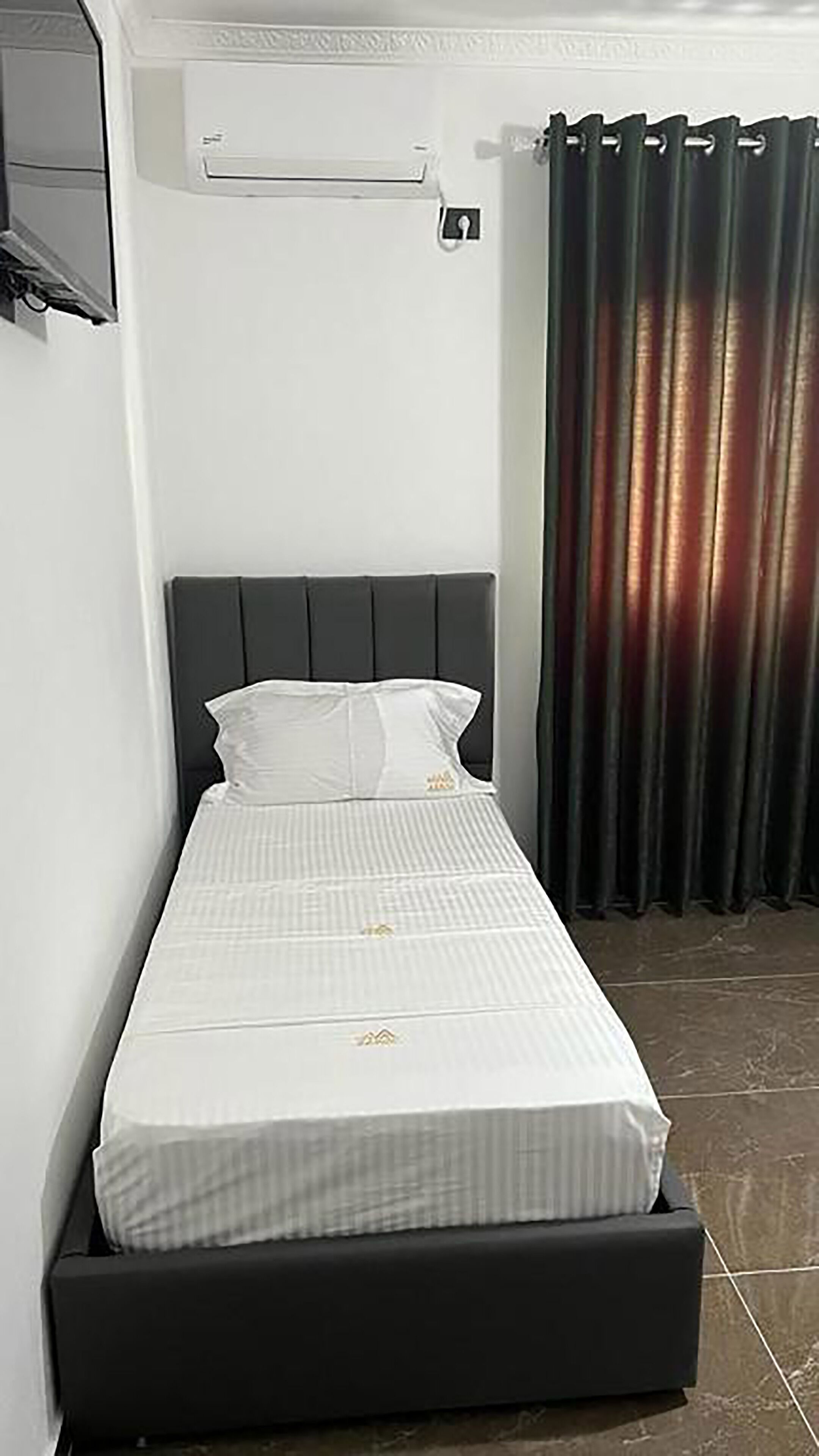 A small, single-bed room with a dark headboard, white bedding, and a curtain on the window.