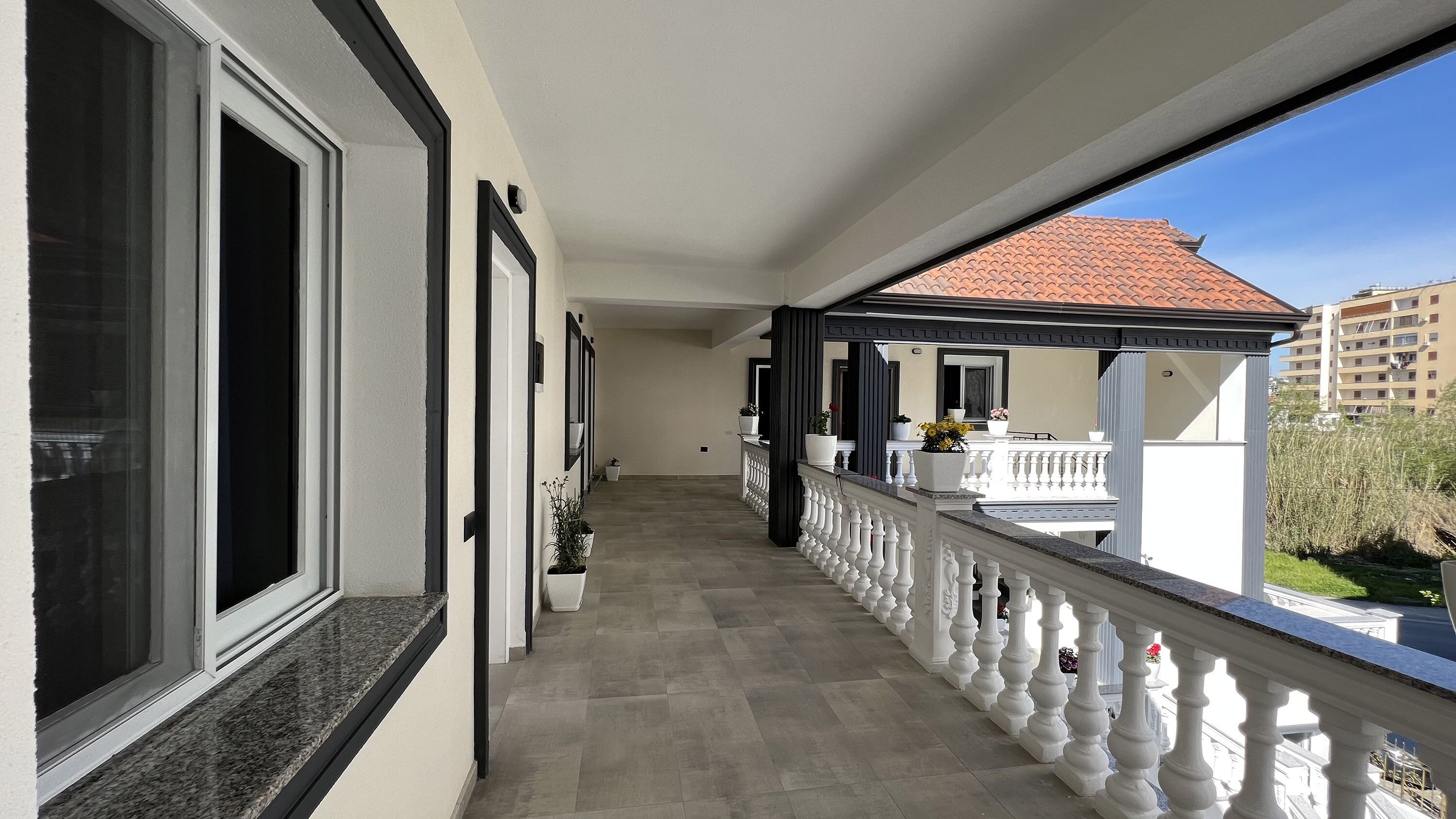 En lang gang med balkon, potteplanter og udsigt til en bygning med rødt tag.