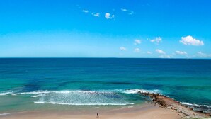 A beach with clear blue water, a rocky shoreline, and a person walking on the sand.