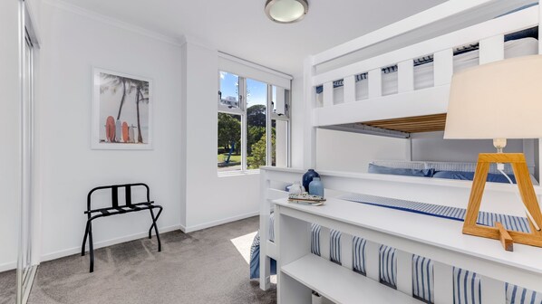 A white room with a bunk bed, a desk, a chair, and a window with a view of trees and buildings. - Kings Edge Unit 1 Kings Beach QLD (Kings Beach)