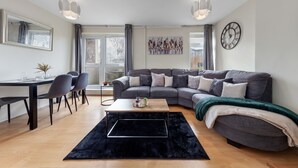 A modern living room with a grey sofa, a black rug, a wooden coffee table, and a dining area with chairs and a table.