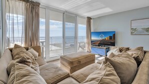 Un salon moderne avec un grand canapé, une table basse et un téléviseur. La pièce est dotée de grandes fenêtres avec des rideaux, offre une vue sur l’océan et porte une enseigne indiquant un service de location.