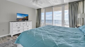 Une chambre avec un grand lit, un ventilateur de plafond, un téléviseur posé sur une commode et une vue sur l’océan grâce à de grandes fenêtres.