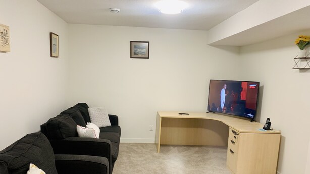 A living room with a dark grey couch, a beige carpet, a wooden desk, and a flat-screen TV. - Centrally located, Cozy two bedroom basement apartment, 10 mins from YYC airport (Calgary)