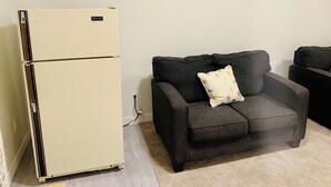 A living room with a beige refrigerator, a dark gray sofa with a decorative pillow, and a small framed picture on the wall. - Centrally located, Cozy two bedroom basement apartment, 10 mins from YYC airport (Calgary)