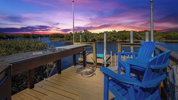 A wooden deck with blue chairs and a metal railing overlooking a body of water with a sunset in the background.