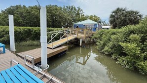 A wooden dock leading to a house with a blue roof, surrounded by greenery and a calm water body.