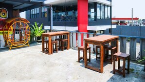 An outdoor seating area with wooden tables and chairs, a swing set, and a building with large windows in the background. - SPOT ON 91765 Kaiku Guest House (Tasikmalaya)