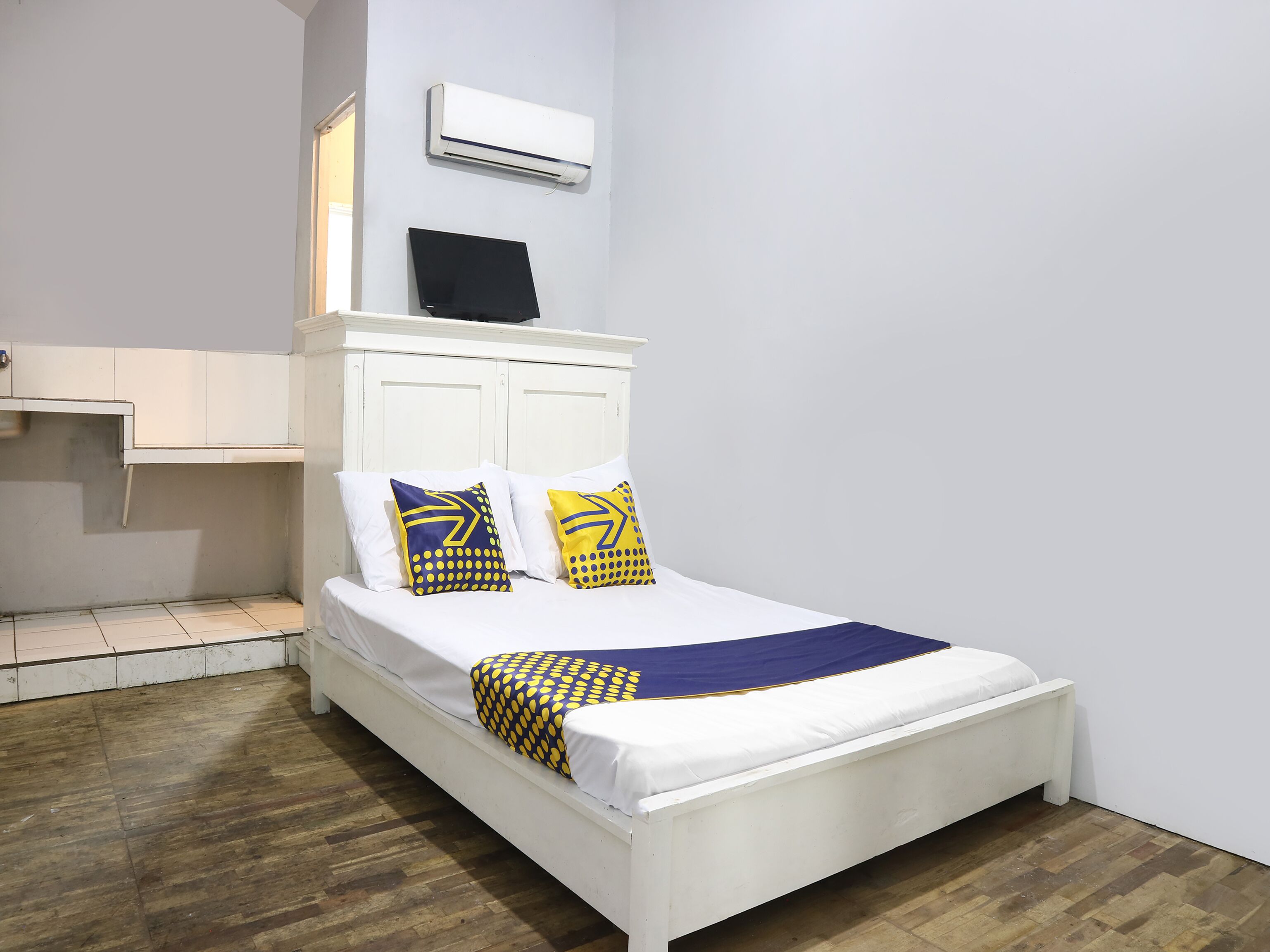 A single bed with white bedding, two pillows with yellow and blue patterns, a white headboard, and an air conditioning unit above.
