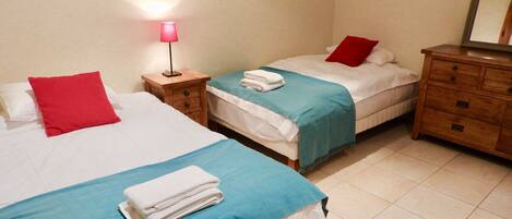 A bedroom with two beds, each with a red pillow, a blue bedspread, and a white blanket. There is a wooden nightstand with a lamp between the beds.