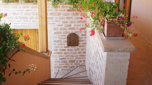Una escalera de madera con un muro de piedra y contraventanas de madera, que conduce a un balcón con plantas y flores en macetas.