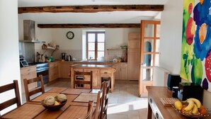 Una cocina con armarios de madera, una mesa de comedor con sillas, un frutero y un cuadro colorido en la pared.
