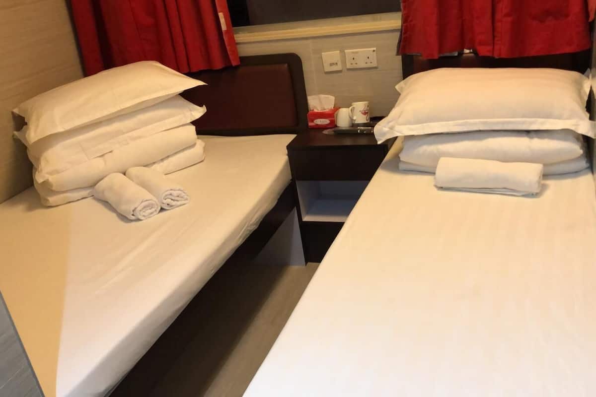 A hotel room with two beds, each with white linens and pillows, a small bedside table with a cup and tissues, and red curtains.