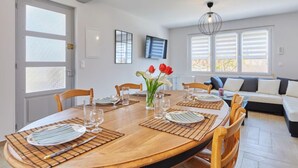 A dining area with a wooden table set for four, surrounded by chairs. There is a sofa with cushions in the background, and a television mounted on the wall. - Janomaine by Interhome (Cabourg)