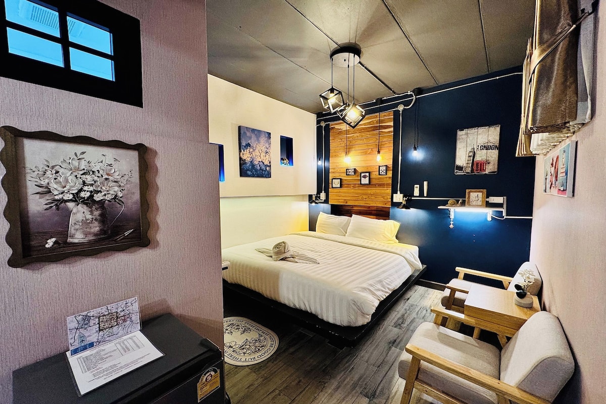 A hotel room with a bed, bedside table, and a small desk with a chair. There are framed pictures on the walls and a window with blinds.