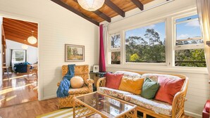 A cozy room with a wicker sofa, a glass coffee table, and a view of trees through large windows. - Cooee Cottage @ Blackheath (Blackheath)